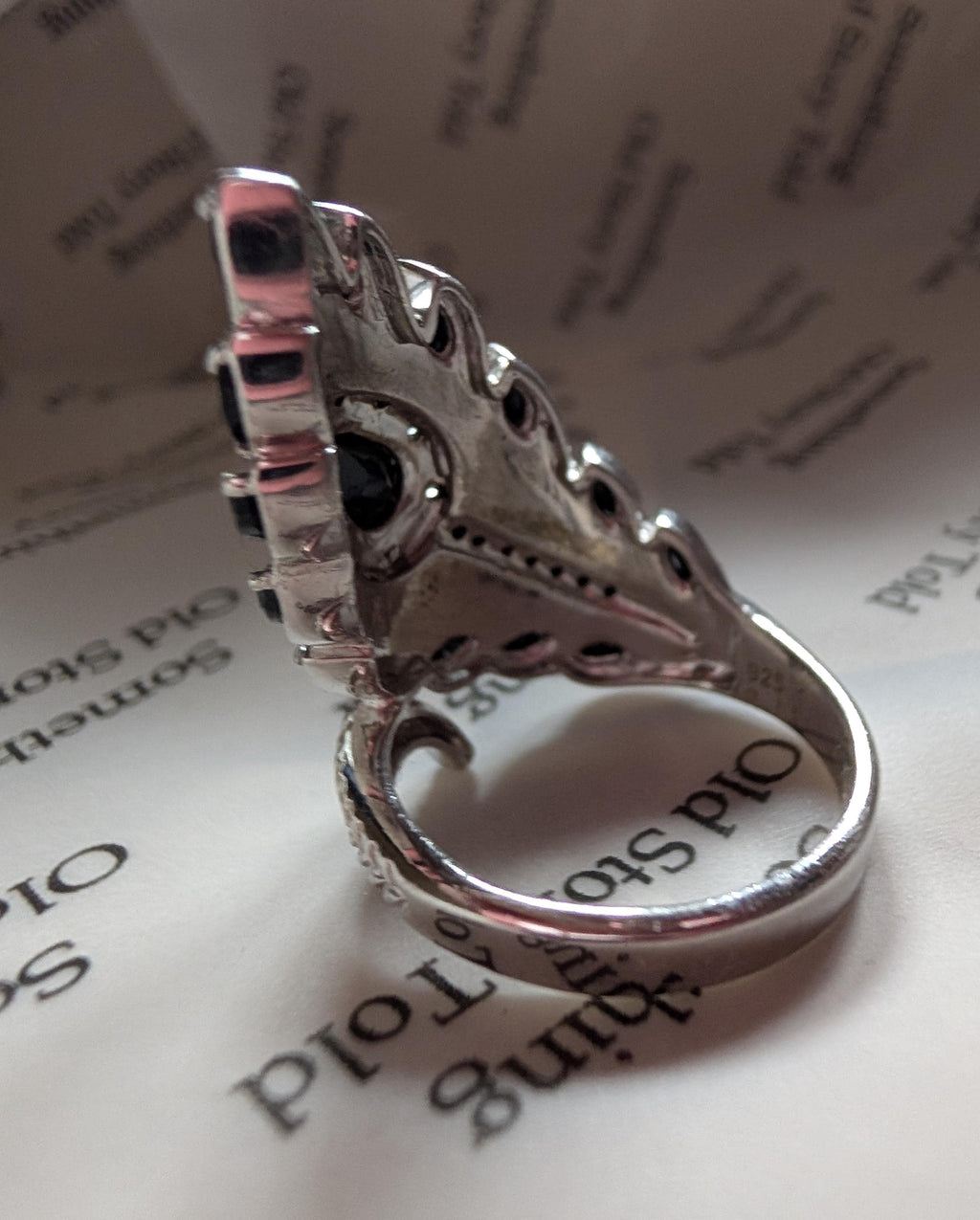 A silver ring with an elaborate design. The ring is set with black gemstones and smaller clear stones. The design is elongated and detailed, with a vintage aesthetic. The band is smooth and polished, reflecting light. The ring is likely made of sterling silver. __etsy:7721462302