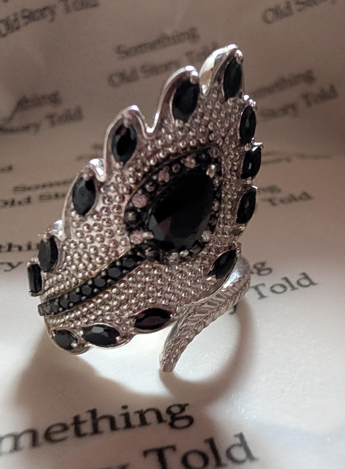 A silver ring with a leaf-shaped design, set with black gemstones and small clear crystals. The ring has a large oval black stone in the centre and smaller stones around the edges. The ring's band has a feather-like detail. __etsy:7721462296