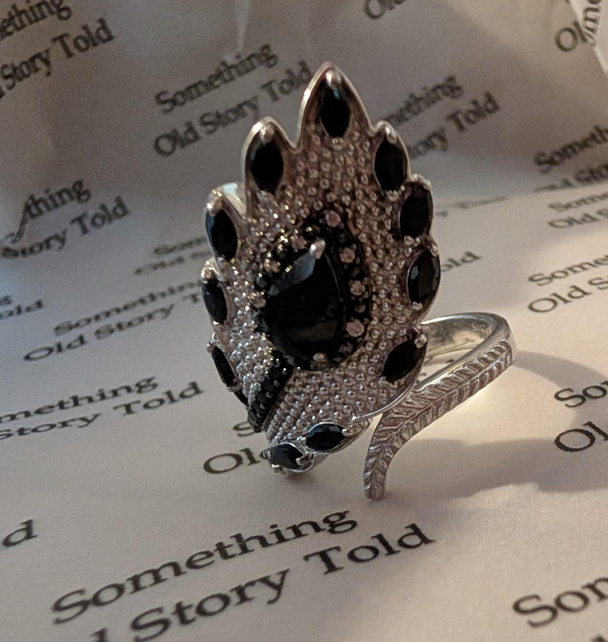 A silver ring with a feather design. The ring has a large, decorative head with black and clear stones. The ring is on paper with the text "Something Old Story Told". __etsy:7769405947
