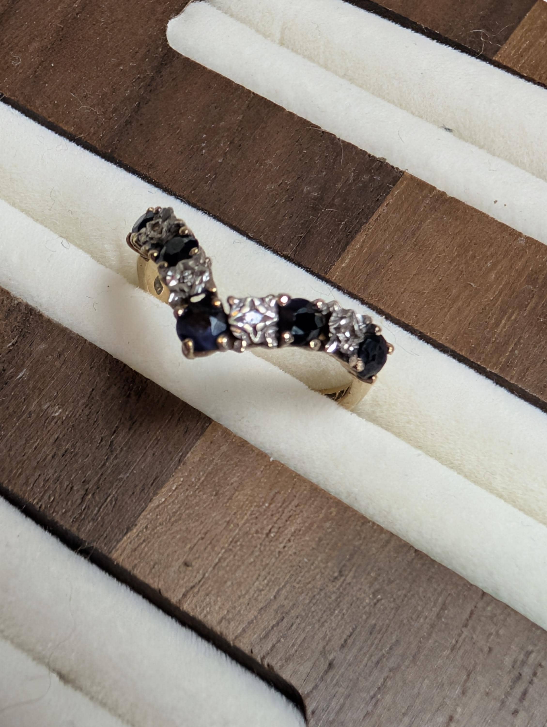 A gold-coloured ring with a V-shaped design. The ring is adorned with alternating dark blue and clear gemstones. The ring is displayed on a white and brown surface. __etsy:7928173017