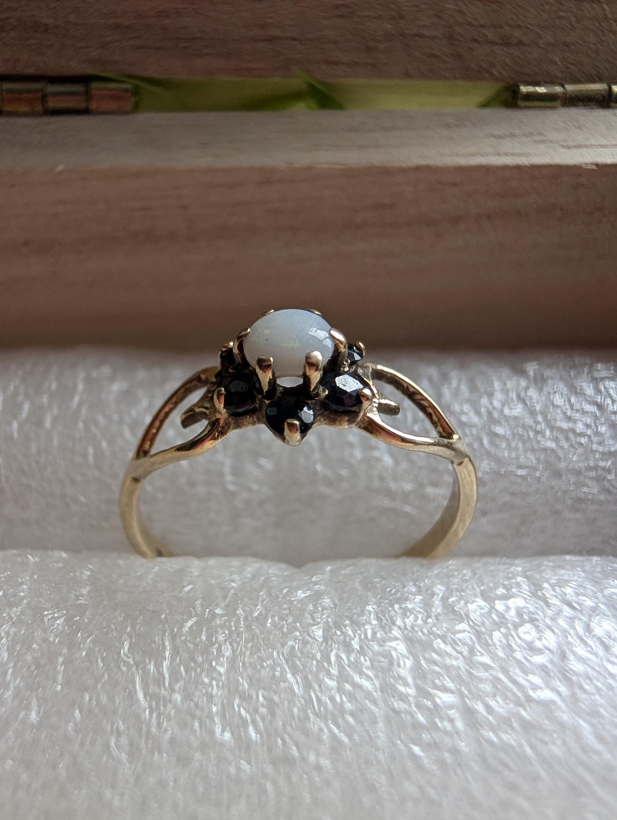 A gold ring with a central oval, milky-white stone, and surrounding dark gemstones. The ring has an openwork design on the band, giving it a vintage look. The ring is set against a soft, white background, creating a contrast. __etsy:7799815783