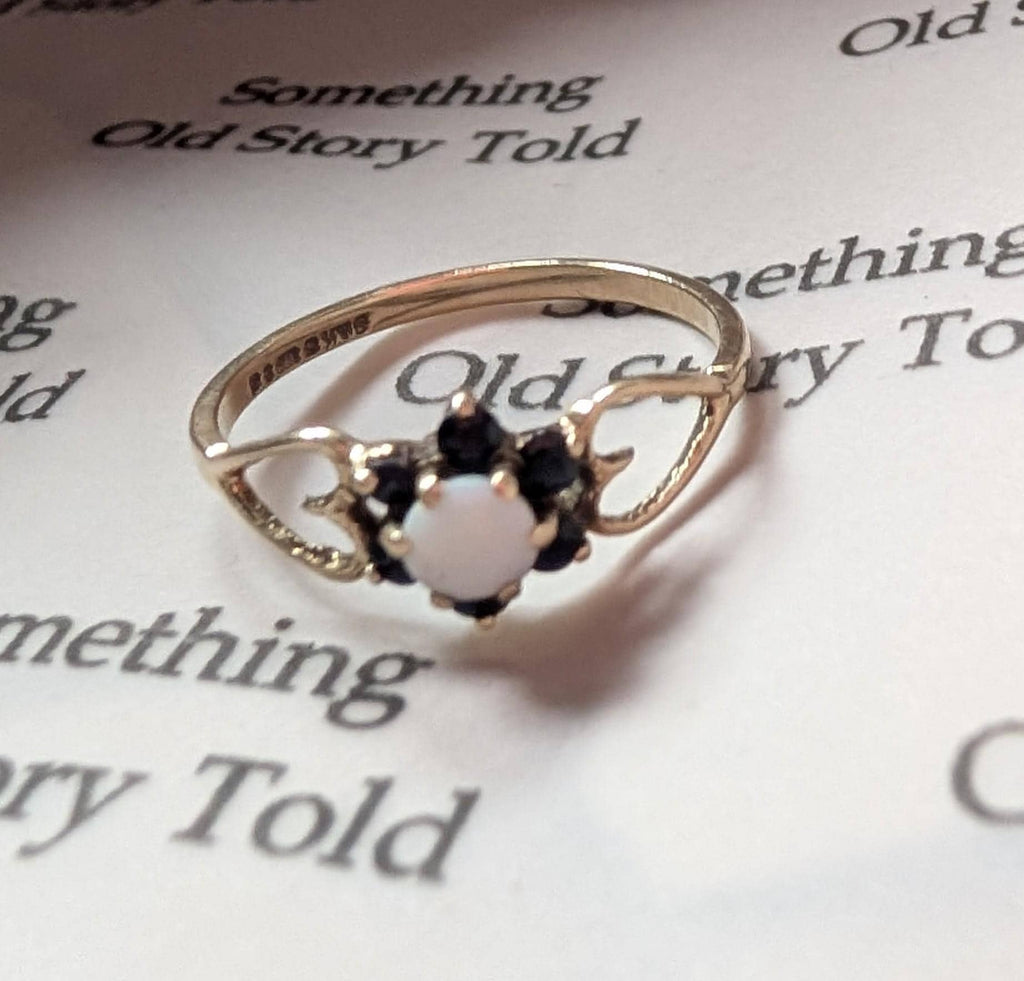 A gold ring with a central white opal and surrounding dark gemstones. The ring has an intricate design with curved details. The ring is set against a backdrop of printed text, with the words "Something Old Story Told". __etsy:7799815731