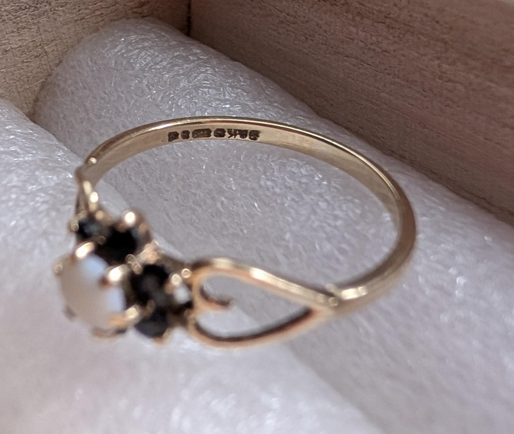 Close-up of a gold ring featuring a central white gemstone and surrounding black stones. The ring has a delicate design with a heart-shaped detail. The ring is set against a white background. __etsy:7751864524