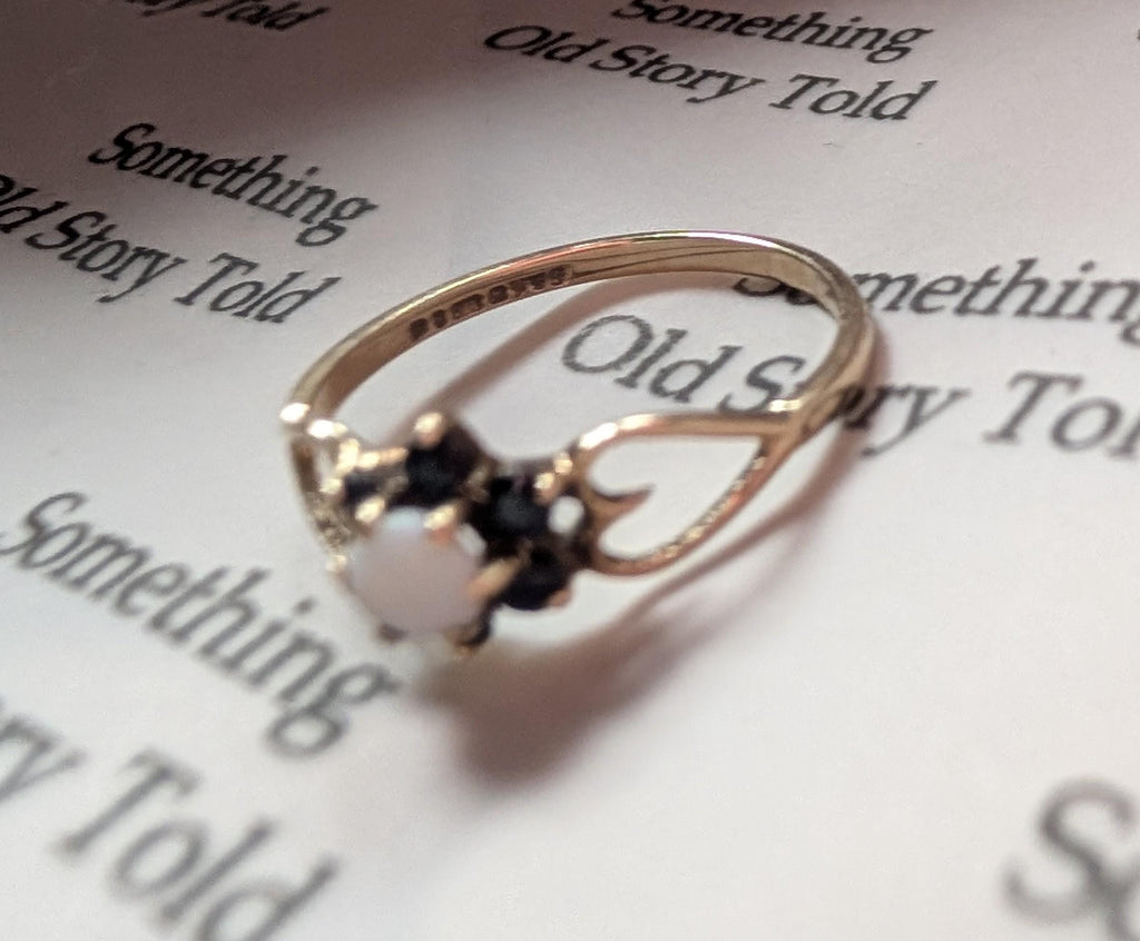 A gold ring featuring a central white gemstone, encircled by smaller dark stones. The ring showcases an intricate design with curved details. The ring rests on a page with the text "Something Old Story Told". __etsy:7751864528