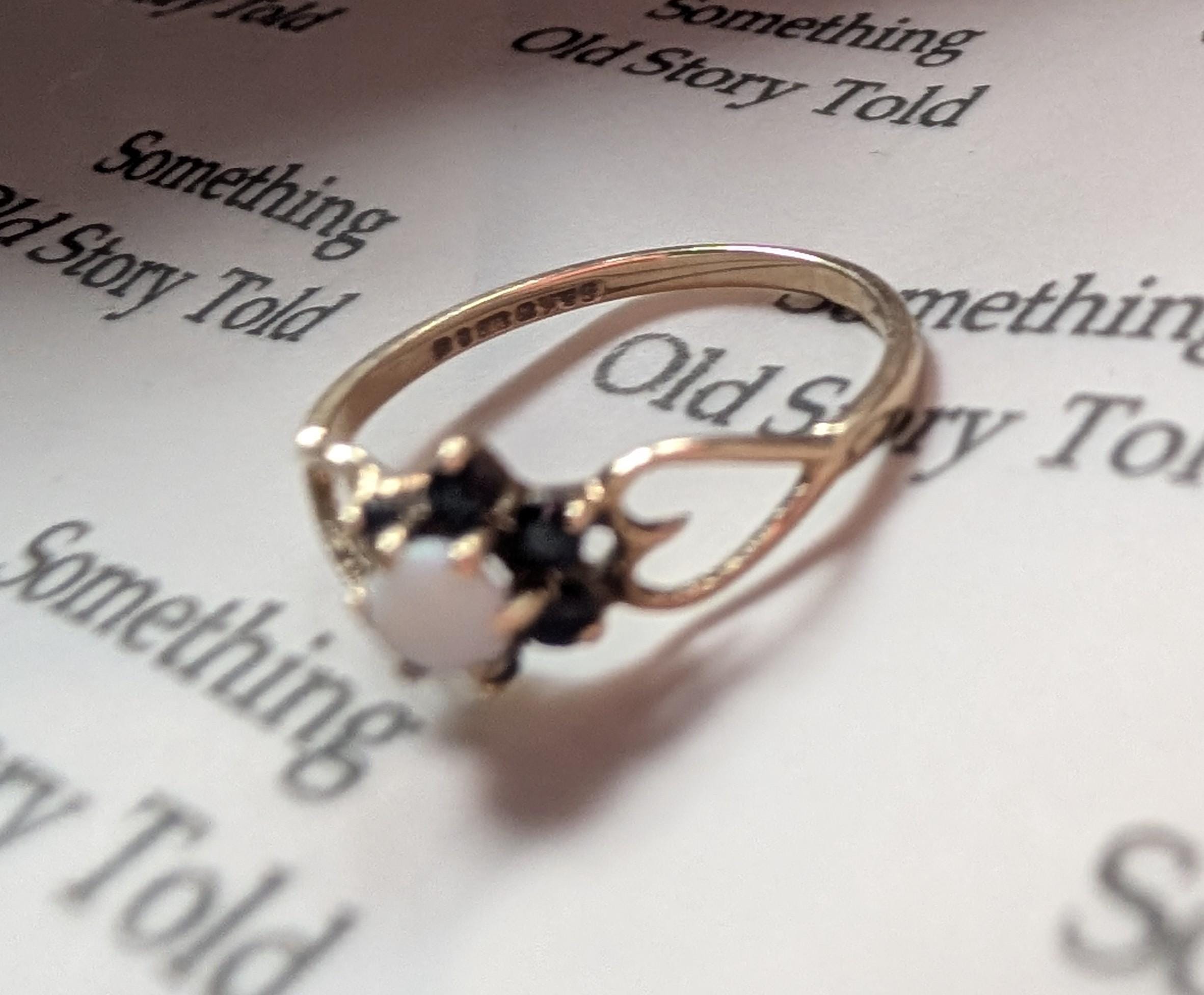 A gold ring featuring a central white gemstone, encircled by smaller dark stones. The ring showcases an intricate design with curved details. The ring rests on a page with the text "Something Old Story Told". __etsy:7751864528