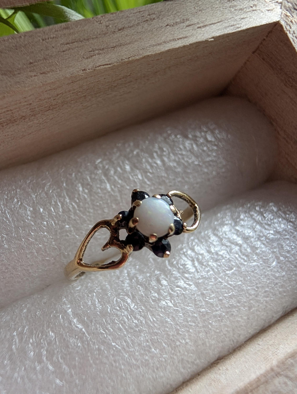 A gold ring with a white opal gemstone at the centre, encircled by dark blue detailing. The ring has a heart-shaped design on the band. The ring is presented in a wooden box. __etsy:7751864582