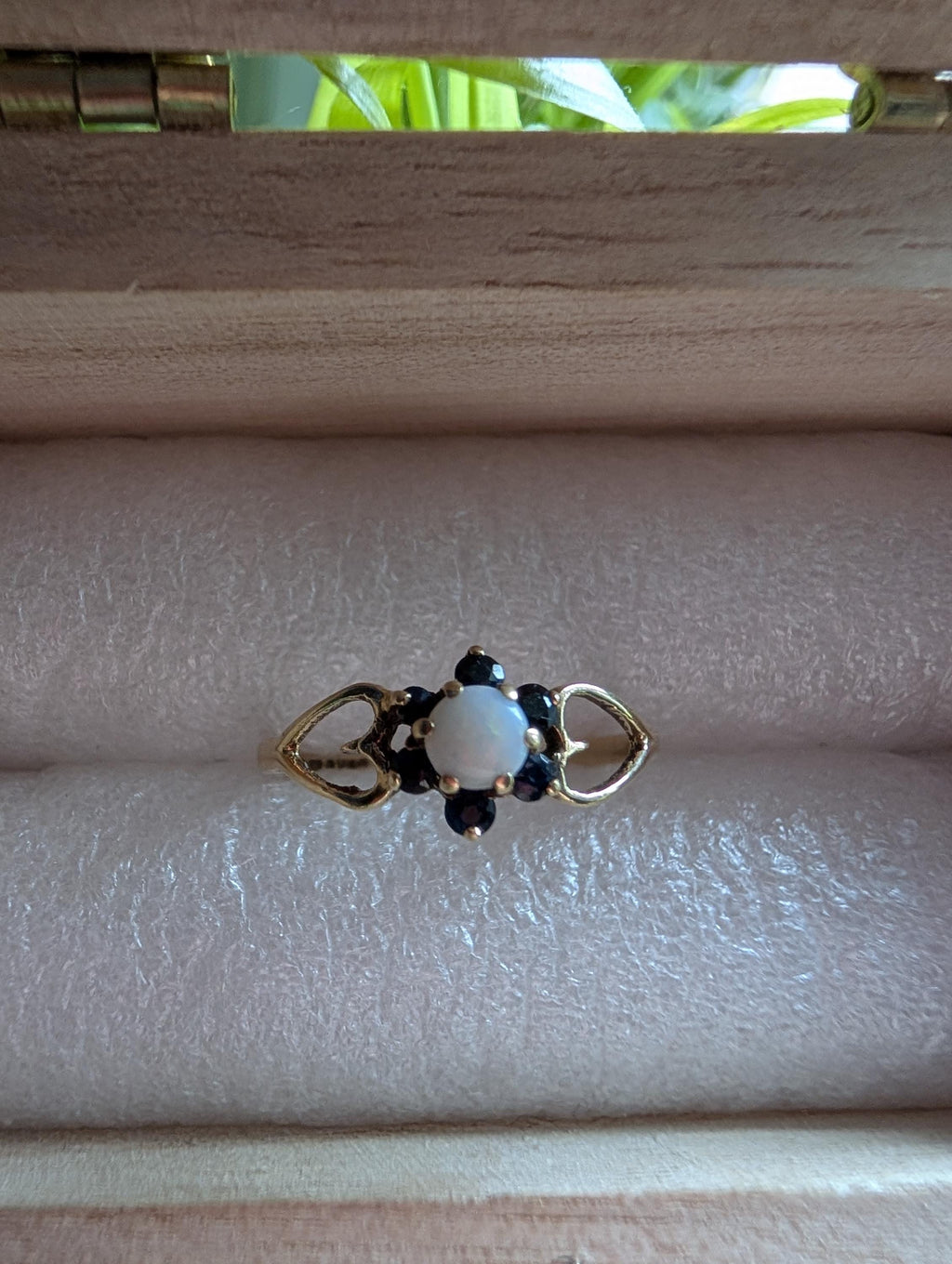 A gold ring featuring a central white opal, encircled by small, dark blue gemstones. The ring showcases a delicate floral design, with heart-shaped cutouts on either side of the central stone. __etsy:7751864568