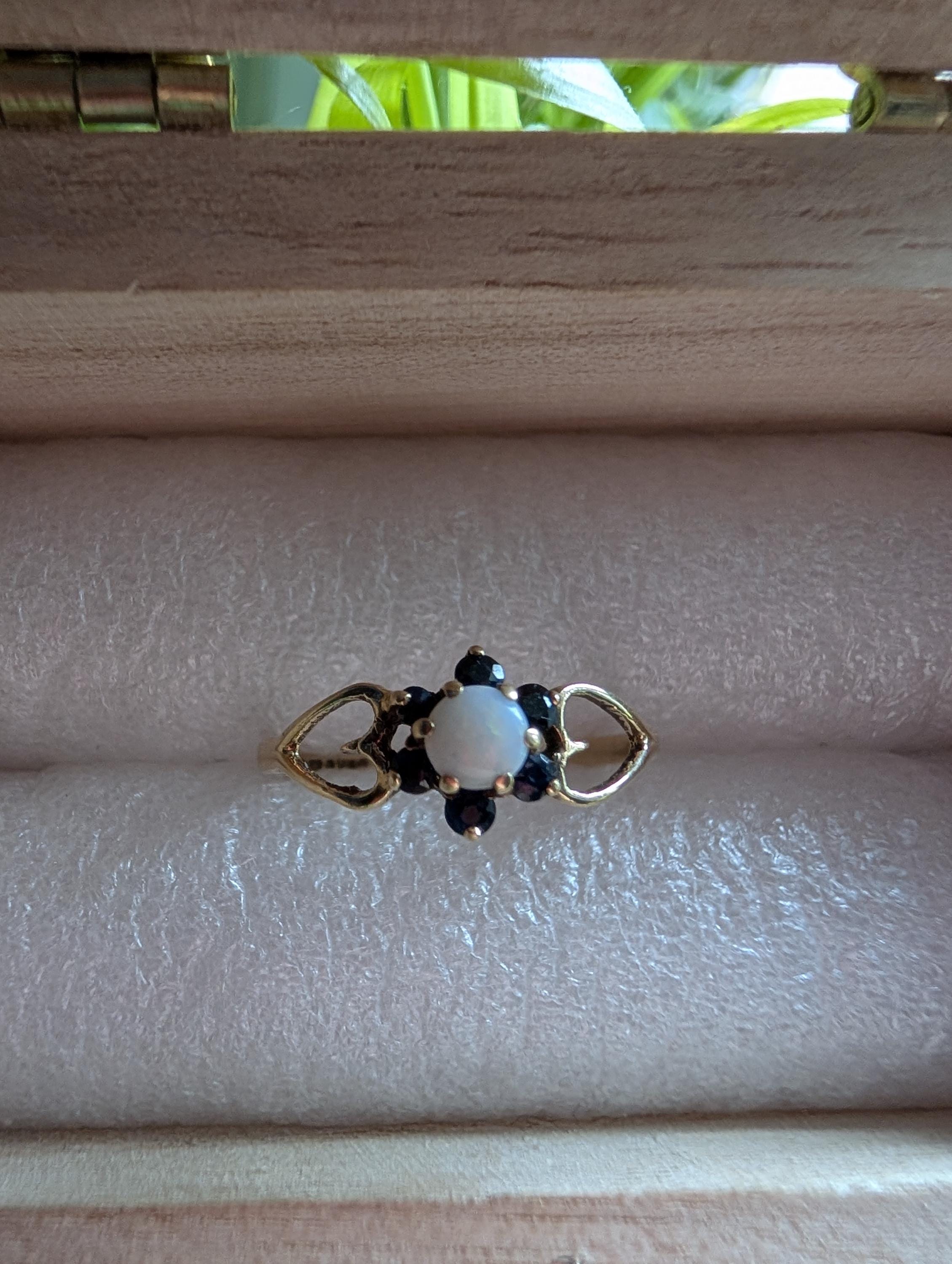 A gold ring featuring a central white opal, encircled by small, dark blue gemstones. The ring showcases a delicate floral design, with heart-shaped cutouts on either side of the central stone. __etsy:7751864568